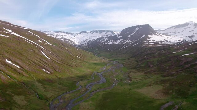 High aerial shot flying over dramatic Icelandic mountain landscape. Steep cliffs and snow, with green grass below.