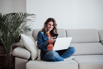 Smiling woman with laptop using smart phone on sofa