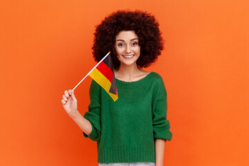 Portrait of attractive positive woman with Afro hairstyle wearing green casual style sweater holding German flag, celebrating Day of German Unity. Indoor studio shot isolated on orange background.