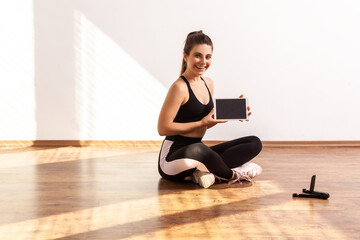 Happy slim woman holding tablet with blank screen, showing sports application for online training, wearing black sports top and tights. Full length studio shot illuminated by sunlight from window.