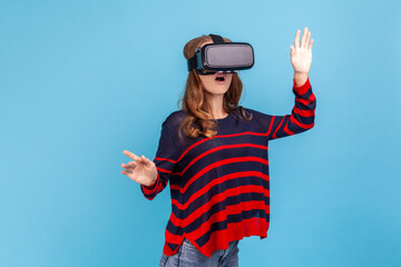 Woman wearing striped casual style sweater and vr headset, playing virtual reality game with shocked face, surprised by innovative technology. Indoor studio shot isolated on blue background.
