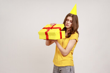 Upset teenager girl opening birthday gift box and looking inside with disappointed expression, unwrapping bad present, wearing yellow T-shirt and birthday cone. Indoor isolated on gray background.