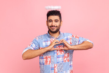 Happy positive handsome man with beard and angelic nimb over head standing and showing heart shape making with hands, looking at camera with love. Indoor studio shot isolated on pink background.