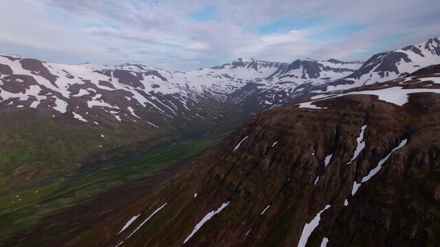 Revolving aerial shot around snowy mountains and green valley below. Shot in Iceland, epic landscape.
