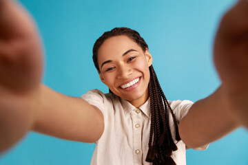 Fototapeta na wymiar Smiling woman with black dreadlocks has happy face expression, stretches arm for making selfie, expresses sincere emotions, point of view photo. Indoor studio shot isolated on blue background.