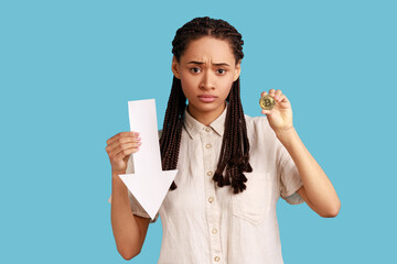 Portrait of dissatisfied woman with dreadlocks showing gold bitcoin and arrow pointing down,...