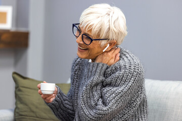 An adult beautiful woman in a grey sweater and glasses sitting at home on a sofa with a mug in her...