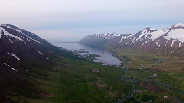 Aerial shot flying towards coastal town in a fjord in Iceland. Snow covered mountains surrounding.