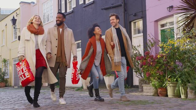Two multi-cultural couples arm in arm as they walk along cobbled mews street on visit to city in autumn or winter carrying sale shopping bags - shot in slow motion