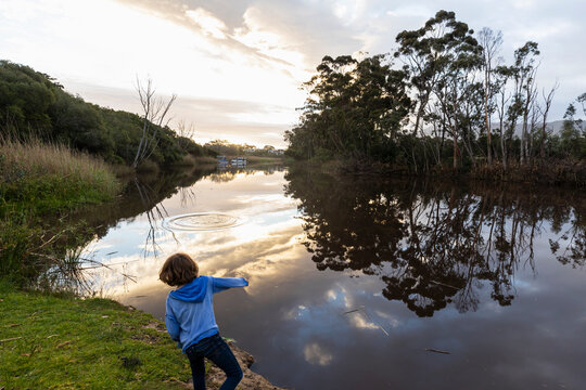 A young boy standing by a river at dusk, sky reflections in the flat calm water