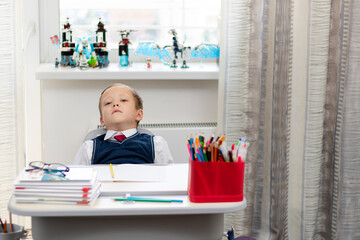 A cute boy first grader in a school uniform and a tie at home during a pandemic does homework while sitting at his desk. Selective focus. Close-up. Portrait