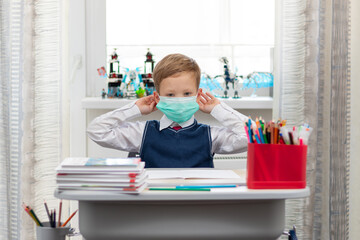 A cute first grader boy in a school uniform at his desk wearing a protective medical mask during...