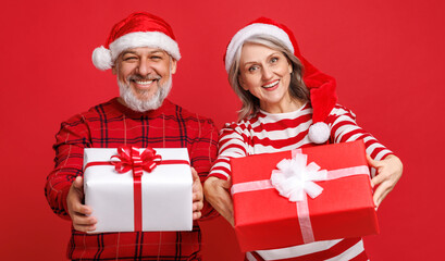 cheerful senior family couple in love  dressed in red Santa hats  holds a Christmas gifts and laughing a colored red background
