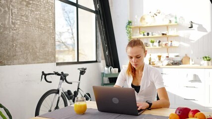 Young female in sportswear is typing on notebook, sitting at table in kitchen at home. Indoor cycle trainer nearby. Slow motion - Powered by Adobe