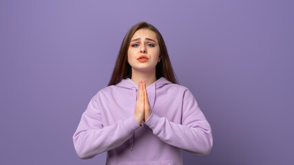 Young woman prayer hopes for better, keeps palms pressed together, looks with hopeful expression, has pleasant appearance, standing in trendy hoodie over purple background