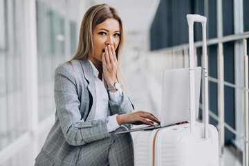 Young woman with travel bag, booking a flight on laptop in airport