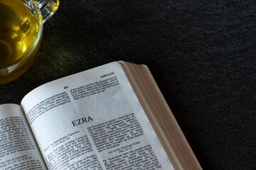 Open Holy Bible Book with a cup of cozy hot tea with yellow color on a black granite table. Studying Old Testament Ezra Scriptures. Christian biblical concept. of obedience to God and restoration. - obrazy, fototapety, plakaty