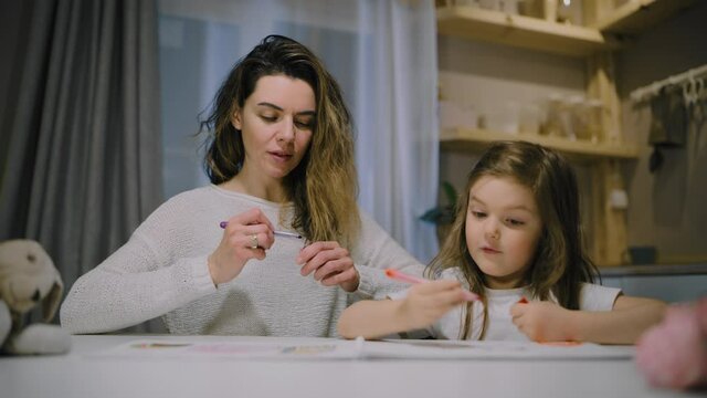 A mother and a small daughter draw drawings on paper in the kitchen with markers in the evening.