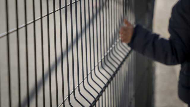 BLurred Out Of Focus Iamge Of Boy Walking By Ht Emetal Fence And Touching Net With Hand