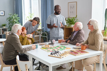 Group of senior people playing board game at the table and talking to medical staff in the room of...