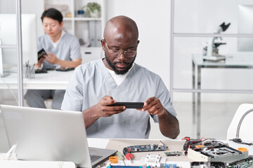 African repairman with smartphone photographing circuit board by workplace against busy female...