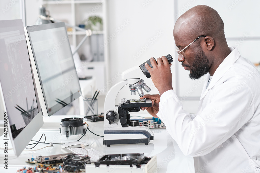Poster Side view of young African scientist in whitecoat studying microchip characteristics in microscope