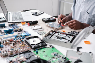 Hands of young African man repairing circuit board with soldering iron while sitting by desk in...