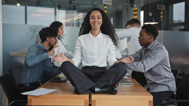Young girl meditate in lotus position sitting on table in office multinational colleagues quarreling in background throwing papers conflict at workplace attractive hispanic woman smiling shows ok