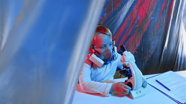 Little boy in potective mask and eyeglasses is sitting in a research lab and looking to the microscope.Little sientist concept