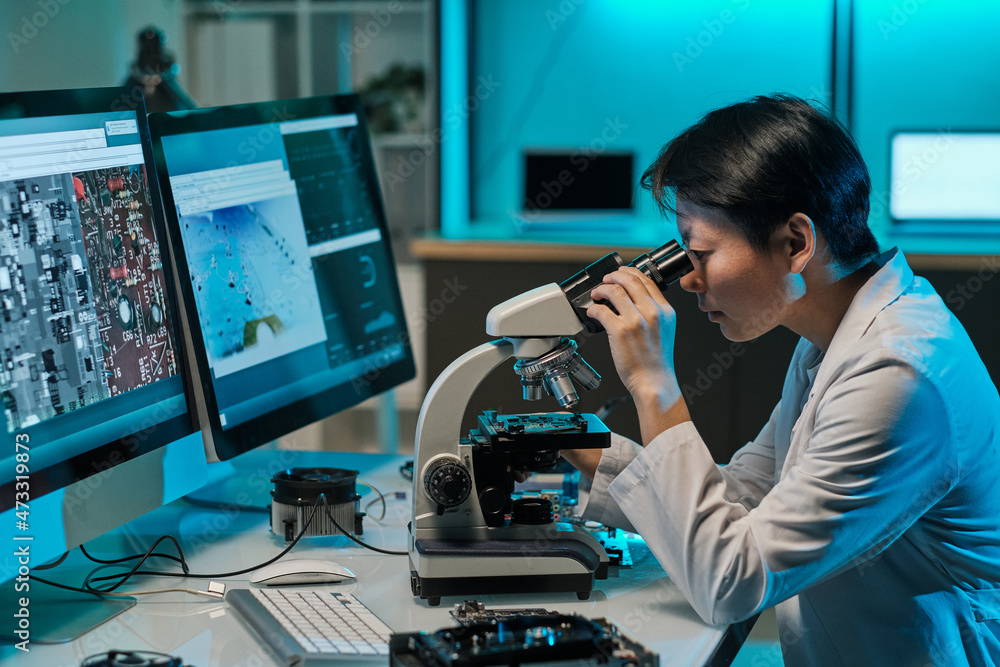 Poster asian female scientist looking in microscope during scientific investigation in front of two compute