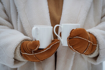 Hello winter, cozy winter mood. A girl in a white fur coat and warm mittens holds two tea cups in her hands, a warm drink to warm up on a frosty day