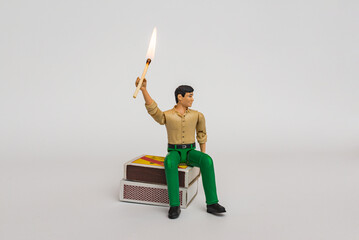 Miniature toy businessman figure sitting on a matchbox holding burning mach stick, on white gray background.