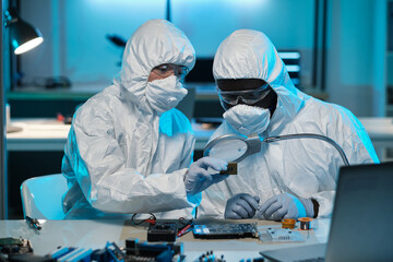 Young Asian woman in coveralls helping colleague with repairment of microchip of electronic device