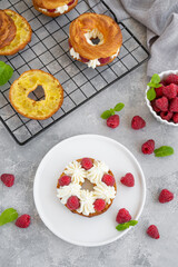 Choux pastries. Choux rings with cream of cream cheese or cottage cheese and fresh raspberries, dusted with powdered sugar on a white plate on a gray concrete background.