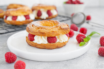 Choux pastries. Choux rings with cream of cream cheese or cottage cheese and fresh raspberries, dusted with powdered sugar on a white plate on a gray concrete background.