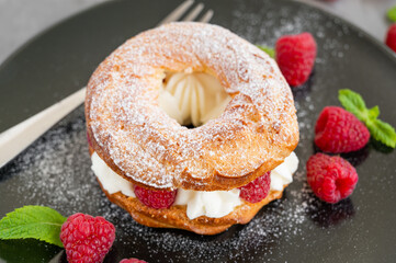 Choux pastries. Choux rings with cream of cream cheese or cottage cheese and fresh raspberries, dusted with powdered sugar on a black plate on a gray concrete background.