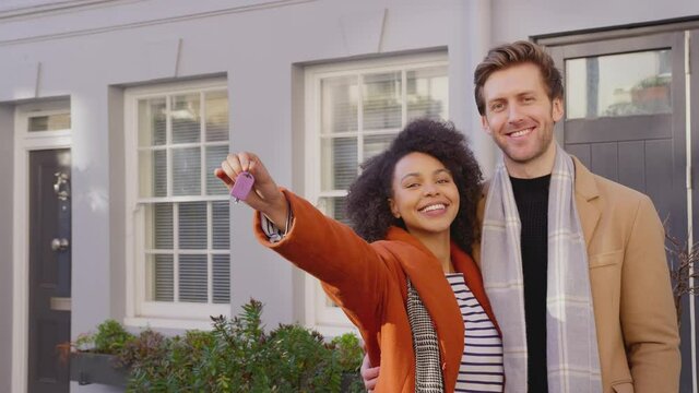 Portrait of multi cultural couple outside house on moving day holding keys to new home in fall or winter - shot in slow motion