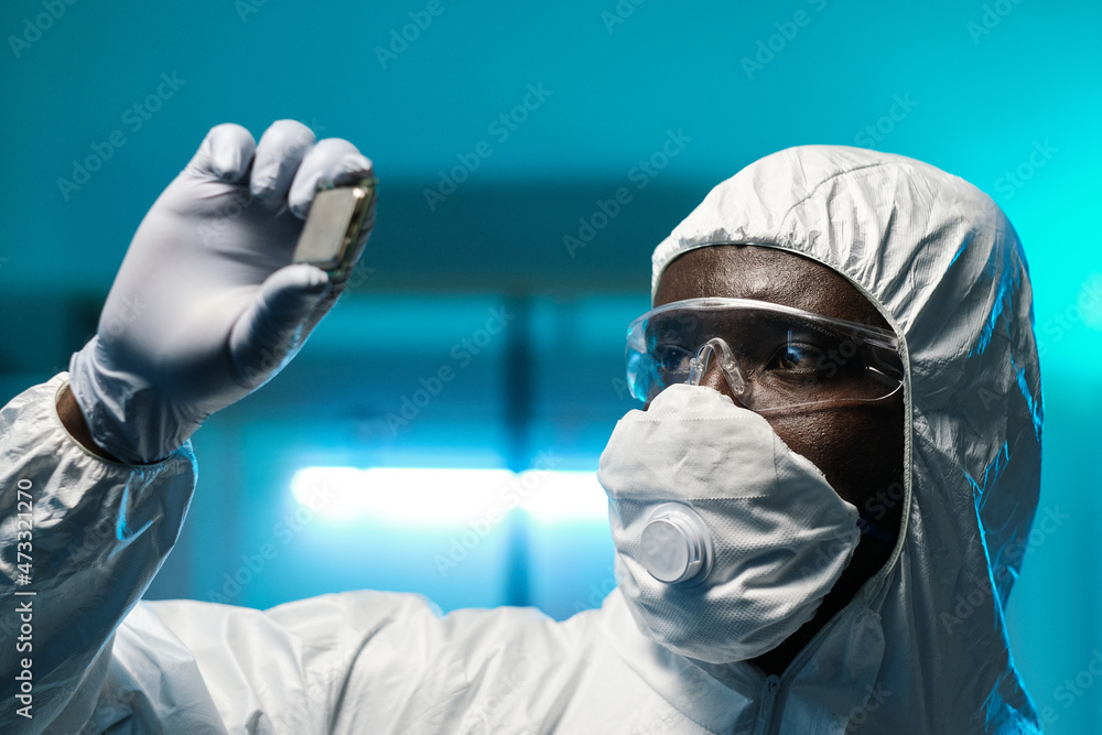 Poster gloved african scientist in protective coveralls, eyeglasses and respirator holding microchip while 