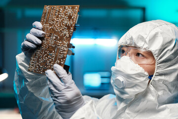 Young repairperson in protective coveralls, gloves, respirator and eyeglasses holding motherboard