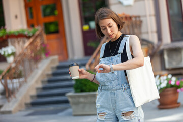 Stylish young woman in denim overalls holding a cup of coffee and looking at the clock in the city