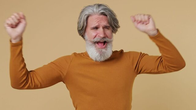 Fun elderly gray-haired mustache bearded man 55 years old wears brown shirt dance waving rising expressive gesticulating hands have fun enjoy isolated on plain pastel beige background studio portrait