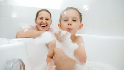 Positive smiling boy with mother playing and having fun with soap foam in bath. Concept of family time, children development and fun at home