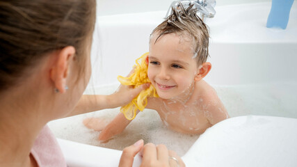 Happy smiling boy looking at mother while she is washing him with soap and sponge in bathroom....