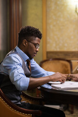 Serious young African male lawayer looking through juridical documents while sitting by table in boardroom