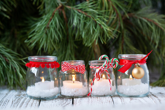 Four candles in glass jars with fir on holiday background. Cozy handmade holiday home decor: glass jar with candle decorated with red ribbon. Christmas decorations.