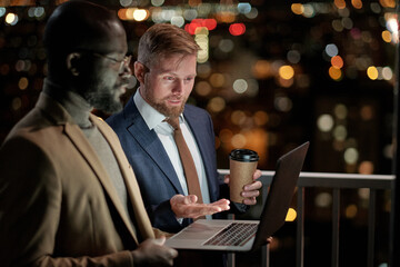 One of two business partners explaining point of presentation while looking at laptop screen at meeting