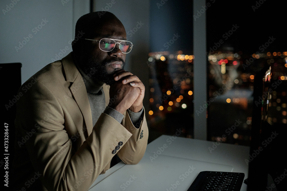 Poster serious businessman in formalwear looking at computer screen while sitting by desk in office at nigh