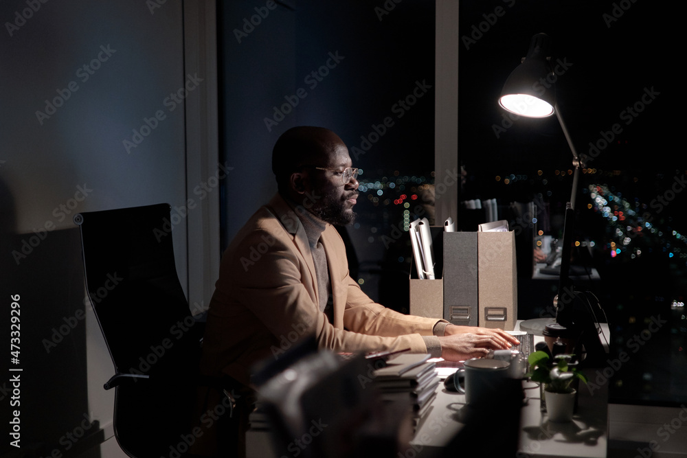 Poster Busy young African man typing on computer keyboard and looking at screen while working late in office