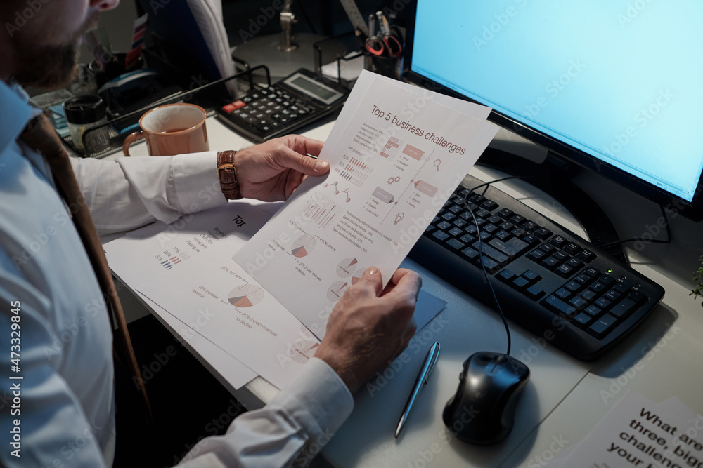 Poster hands of young businessman holding financial documents while sitting by desk in front of computer mo