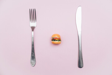 Miniature burger, fork and knife on a pastel pink background. Minimalistic food concept. Creative idea. Diet.